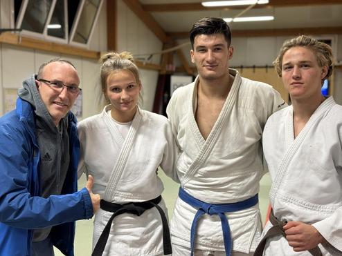 Vier Personen in Judo-Anzügen stehen gemeinsam in einem Trainingsraum, lächelnd, mit einem Trainer links im Bild.