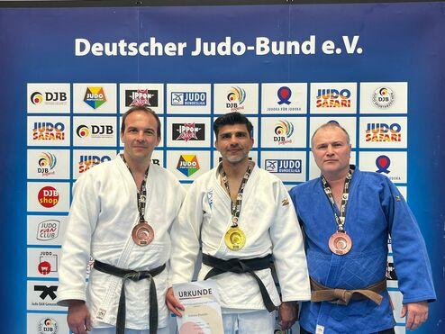 Drei Judokas in Gi mit Medaillen stehen vor einem Werbetransparent des Deutschen Judo-Bunds e.V. Halten eine Urkunde.