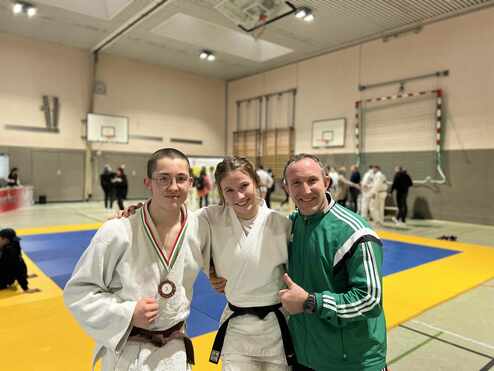 Drei Personen in Judokleidung posieren lächelnd auf einer Wettkampfmatte, eine trägt eine Medaille um den Hals.