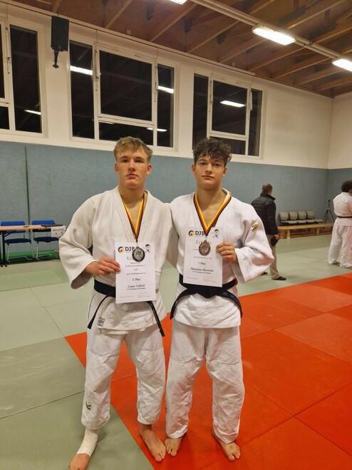 Zwei Judoka in weißen Anzügen posieren mit Medaillen und Urkunden in einer Trainingshalle auf einer Tatami.