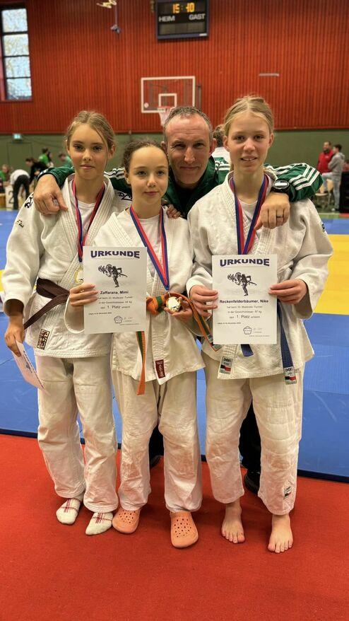 Drei Judokas in weißen Anzügen zeigen stolz ihre Urkunden und Medaillen, umarmt von einem Trainer in grünem Outfit.