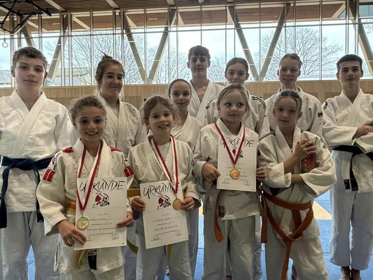 Gruppe junger Judoka in weißen Anzügen, einige halten Medaillen und Urkunden, auf einer blauen Judomatte.