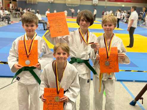 Vier Jungen in Judoanzügen halten Medaillen und Urkunden in einer Turnhalle mit blauen und gelben Matten.