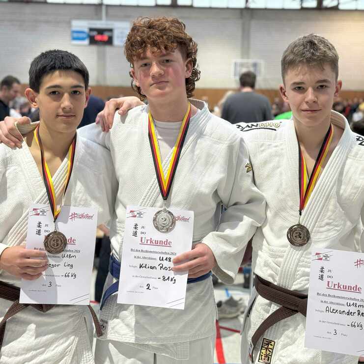 Drei Jugendliche in Judo-Anzügen halten Medaillen und Urkunden nach einem Wettkampf in einer Sporthalle.