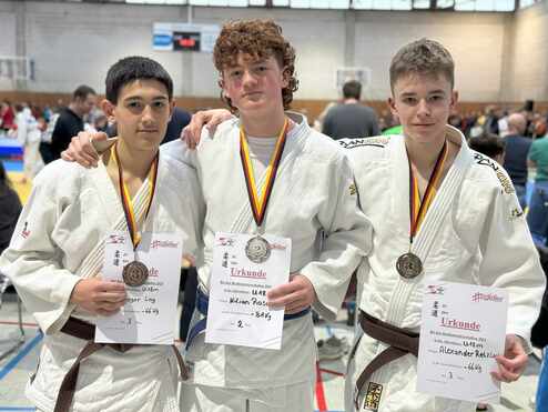 Drei Jugendliche in Judo-Anzügen halten Medaillen und Urkunden nach einem Wettkampf in einer Sporthalle.