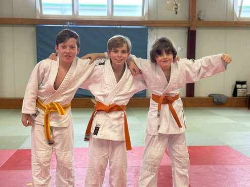Drei Kinder in weißem Judoanzug mit verschiedenen Gürtelfarben stehen in einer Judohalle und lächeln in die Kamera.