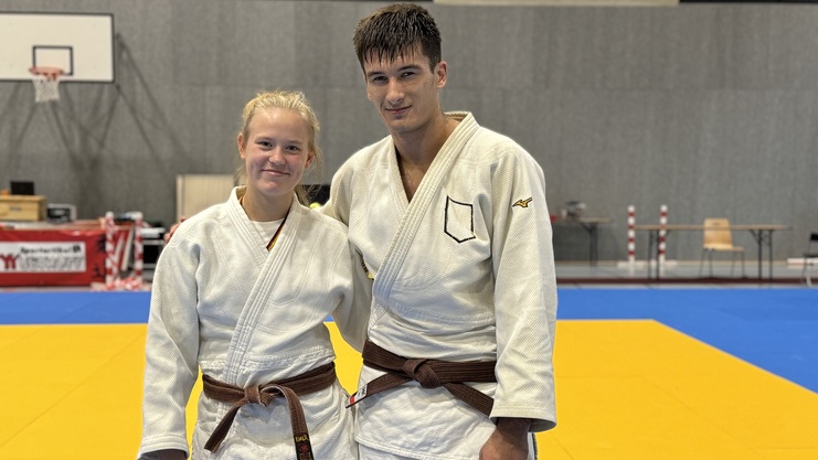 Zwei Judokas in wei&szlig;en Anz&uuml;gen und braunen G&uuml;rteln l&auml;cheln auf einer Judomatte in einer Sporthalle.