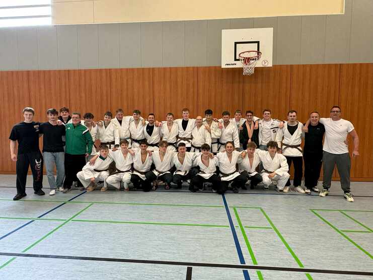 Gruppe von Judokas in weißen Gi mit Schwarz- und Braungurten, posierend in einer Sporthalle vor einer Holzwand.