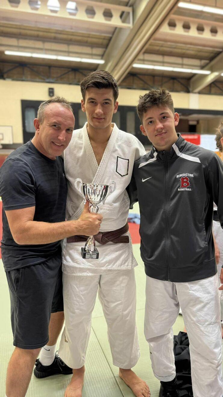 Drei junge Männer stehen auf einer Judo-Matte, einer hält einen Pokal, alle lächeln in die Kamera.