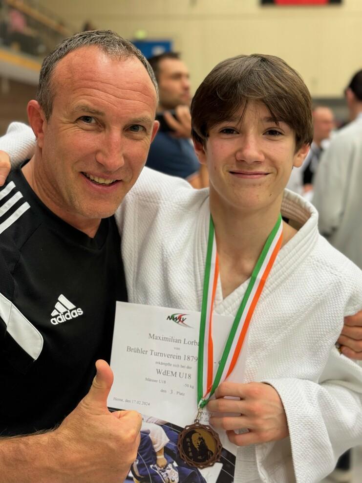 Zwei Männer lächeln, der Junge hält eine Urkunde und eine Medaille nach einem Judowettkampf.