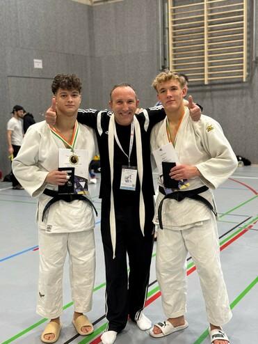 Drei Judoka posieren nach einem Wettkampf in Sportbekleidung, zwei halten Medaillen, einer zeigt Daumen nach oben.