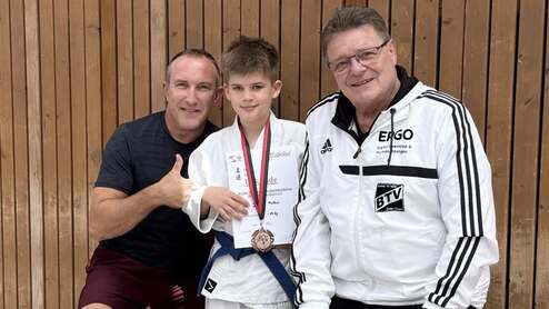 Junge in Karate-Gi hält Medaille und Urkunde, posiert mit zwei Erwachsenen vor Holzwand.