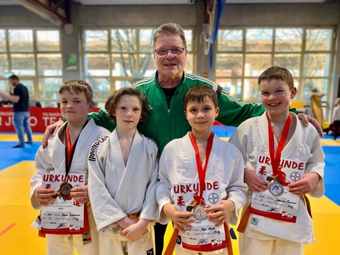 Gruppe von vier Personen in Judogi, lächelnd mit Medaillen und Urkunden, vor einem Judoturnier-Hintergrund.