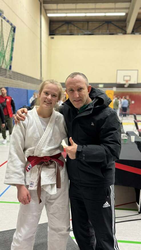 Ein Judo-Athlet in weißem Kimono und braunem Gürtel posiert mit einem Mann in schwarzer Jacke in einer Sporthalle.