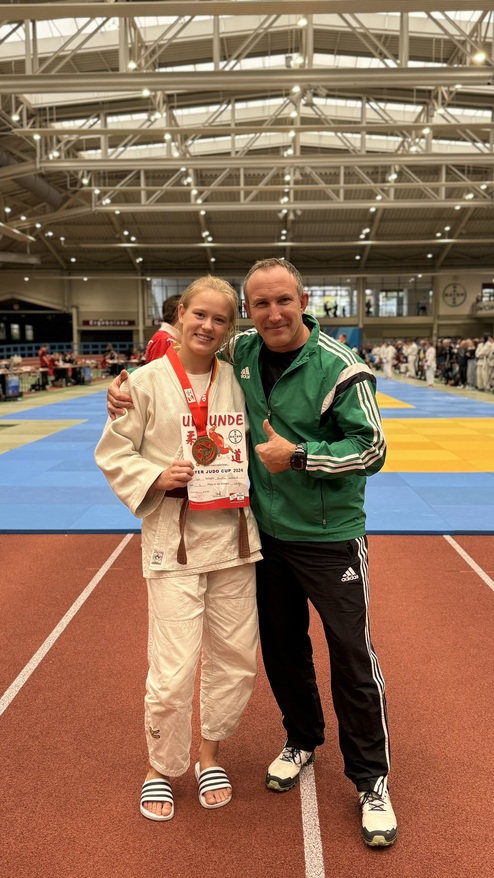Junge Judoka mit Medaille und Urkunde um den Hals, umarmt von einem Trainer auf einer Wettkampfstätte.