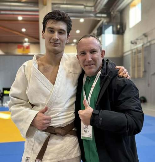Junge im Judoanzug und Mann in Jacke stehen lächelnd mit Daumen hoch auf einer Judomatte.