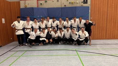 Gruppe von 14 Judoka in weißen Gi-Jacken und braunen Gürteln, lächeln und posieren in einem Sportraum.