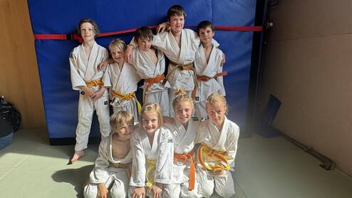 Gruppe von acht Kindern in weißen Judojacken mit farbigen Gürteln, lächelnd vor einer blauen Matte.