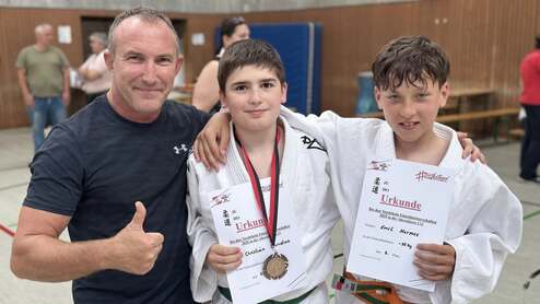 Drei Personen in Judojacken halten Urkunden für einen Wettkampf und zeigen Medaillen in einer Turnhalle.