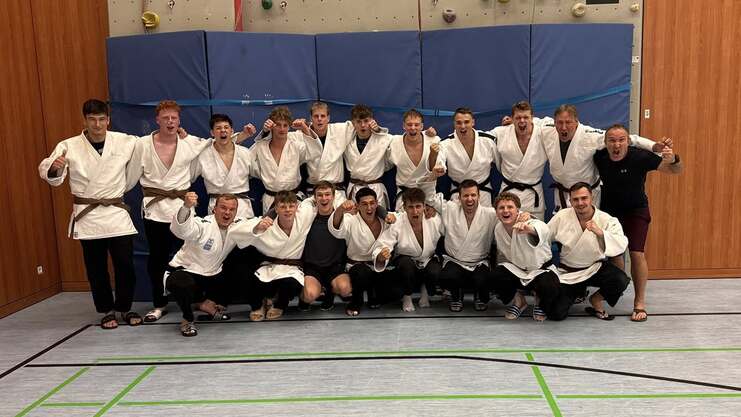 Gruppe von 14 Judoka in weißen Gi-Jacken und braunen Gürteln, lächeln und posieren in einem Sportraum.