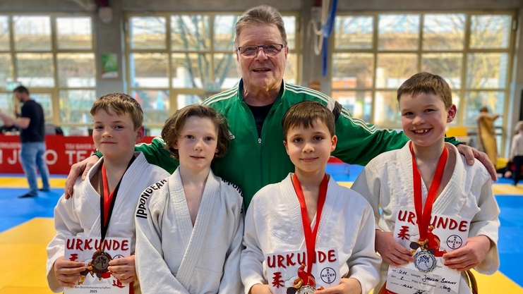 Gruppe von vier Personen in Judogi, l&auml;chelnd mit Medaillen und Urkunden, vor einem Judoturnier-Hintergrund.