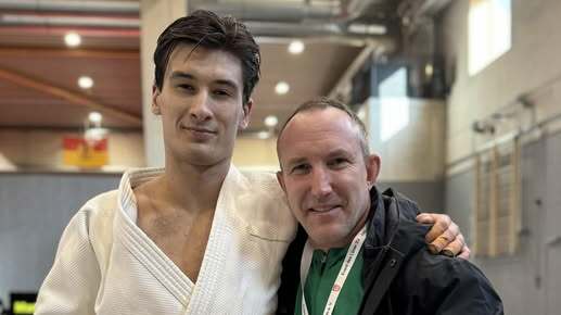 Junge im Judoanzug und Mann in Jacke stehen lächelnd mit Daumen hoch auf einer Judomatte.