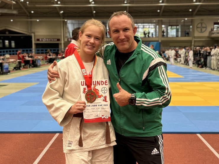 Junge Judoka mit Medaille und Urkunde um den Hals, umarmt von einem Trainer auf einer Wettkampfst&auml;tte.