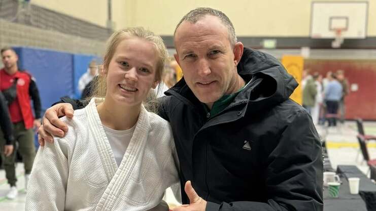 Ein Judo-Athlet in weißem Kimono und braunem Gürtel posiert mit einem Mann in schwarzer Jacke in einer Sporthalle.
