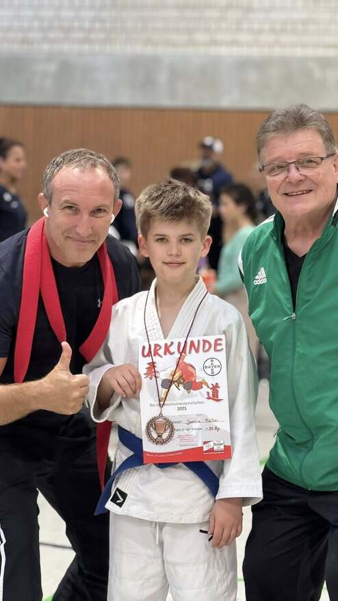 Zwei Erwachsene stehen neben einem Jungen in Judokleidung, der eine Urkunde mit einem Bronze-Podestplatz hält.