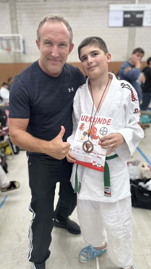 Zwei Personen posieren lächelnd vor einem Hintergrund, der eine Sportveranstaltung zeigt. Ein Junge hält eine Urkunde und Medaille.