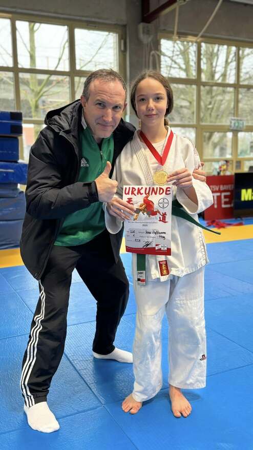 Mädchen in Karate-Gi hält eine Urkunde und Medaille, posiert mit Trainer auf blauen Matten.
