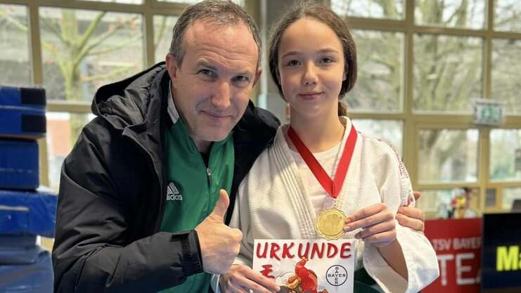 Mädchen in Karate-Gi hält eine Urkunde und Medaille, posiert mit Trainer auf blauen Matten.