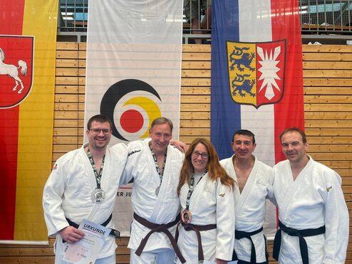 Gruppe von Judokas in weißen Anzügen, die Medaillen halten, vor verschiedenen Flaggen in einer Sporthalle.