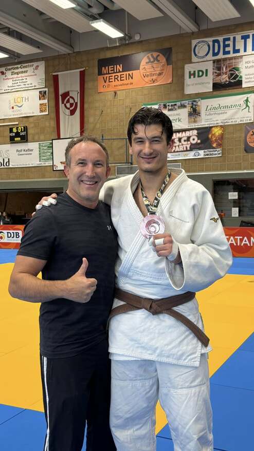 Zwei Männer posieren auf einer Judomatte, einer trägt eine Medaille und einen Judogi, der andere zeigt Daumen hoch.