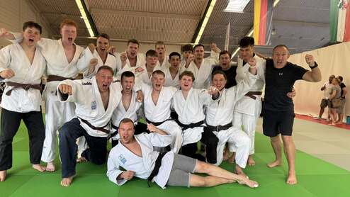Gruppenfoto von 16 Judoka in weißen und schwarzen Judogis, die jubelnd auf einer grünen Matte posieren.