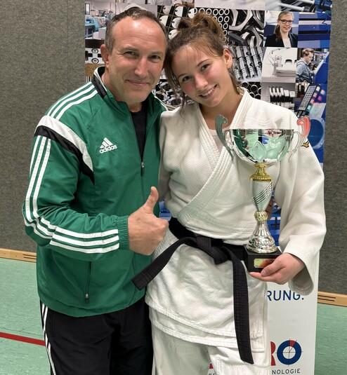 Trainer und Athletin posieren lächelnd mit einer Trophäe nach einem Judo-Wettkampf in einer Sporthalle.