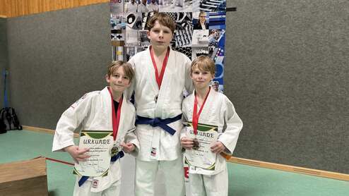 Drei junge Judokas in weißen Judogis mit Medaillen und Urkunden stehen lächelnd vor einer Wand mit Bildern.