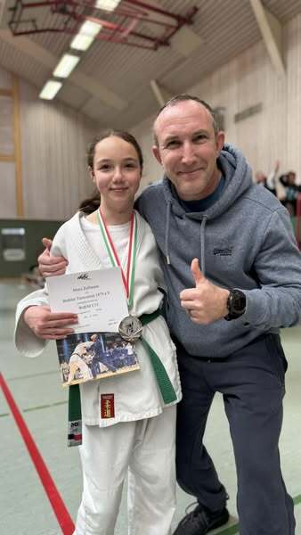 Mädchen in Karate-Gi hält eine Urkunde und Medaille, posiert mit einem Trainer in einer Sporthalle.