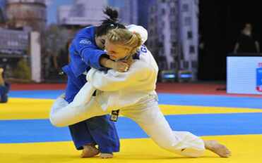 Zwei Judoka k&auml;mpfen auf einer gelben und blauen Matte, eine in blauem, die andere in wei&szlig;em Judogi.