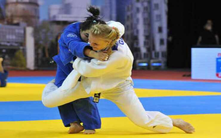Zwei Judoka kämpfen auf einer gelben und blauen Matte, eine in blauem, die andere in weißem Judogi.