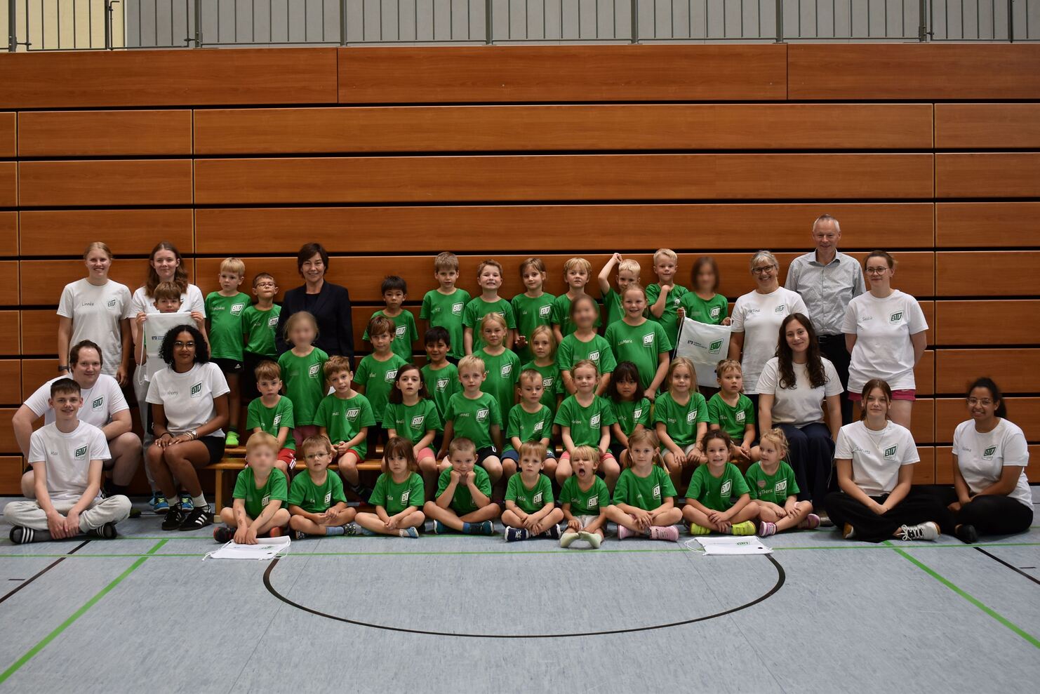 Gruppe von Kindern und Betreuern in Sportkleidung l&auml;chelt zusammen in einer Turnhalle vor Holzverkleidung.