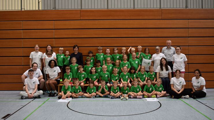 Gruppe von Kindern und Betreuern in Sportkleidung l&auml;chelt zusammen in einer Turnhalle vor Holzverkleidung.