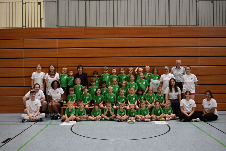 Gruppe von Kindern und Betreuern in Sportkleidung l&auml;chelt zusammen in einer Turnhalle vor Holzverkleidung.