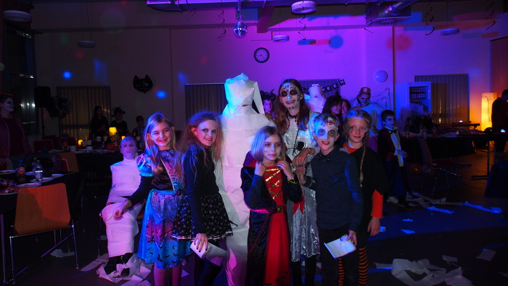 Sechs Kinder in Halloweenkost&uuml;men posieren vor einem mit T&uuml;chern bedeckten Tisch in einem festlich beleuchteten Raum.