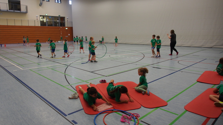 Kinder in gr&uuml;nen T-Shirts spielen in einer Sporthalle mit Reifen und Matten auf dem Boden.