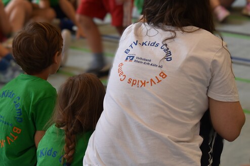Lehrerin mit T-Shirt &bdquo;BTV-Kids Camp&ldquo; sitzt im Kreis von Kindern in gr&uuml;nen T-Shirts in einer Sporthalle.
