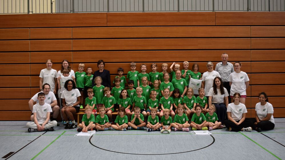 Gruppe von Kindern und Betreuern in Sportkleidung lächelt zusammen in einer Turnhalle vor Holzverkleidung.