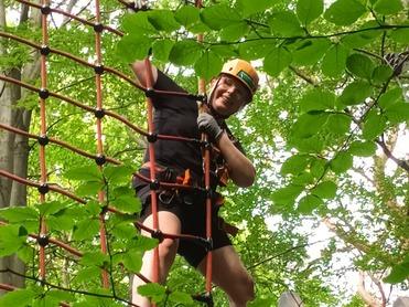 Ein Mann in einem Klettergeschirr steht auf einem Kletterturm und hält sich an einem Netz aus Kletterseilen fest.