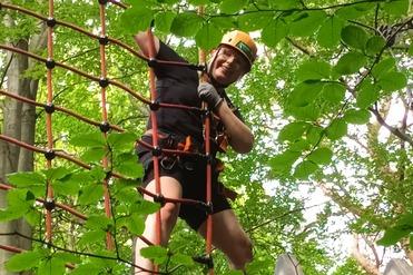 Ein Mann in einem Klettergeschirr steht auf einem Kletterturm und h&auml;lt sich an einem Netz aus Kletterseilen fest.