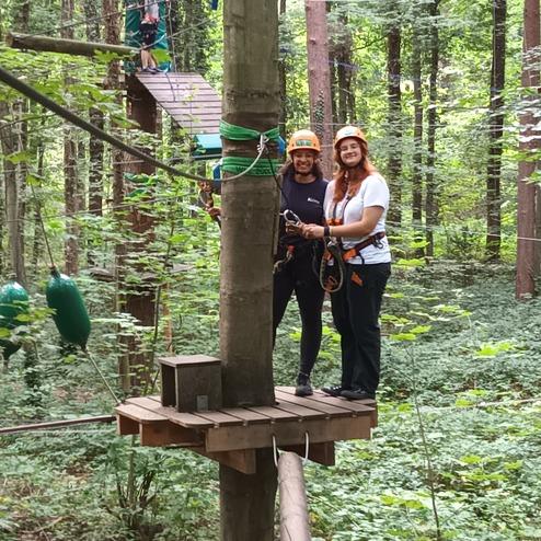 Zwei Personen in Sicherheitsgeschirr und -helmen stehen auf einer Plattform im Wald, w&auml;hrend ein dritter Teilnehmer sich vorbereitet.